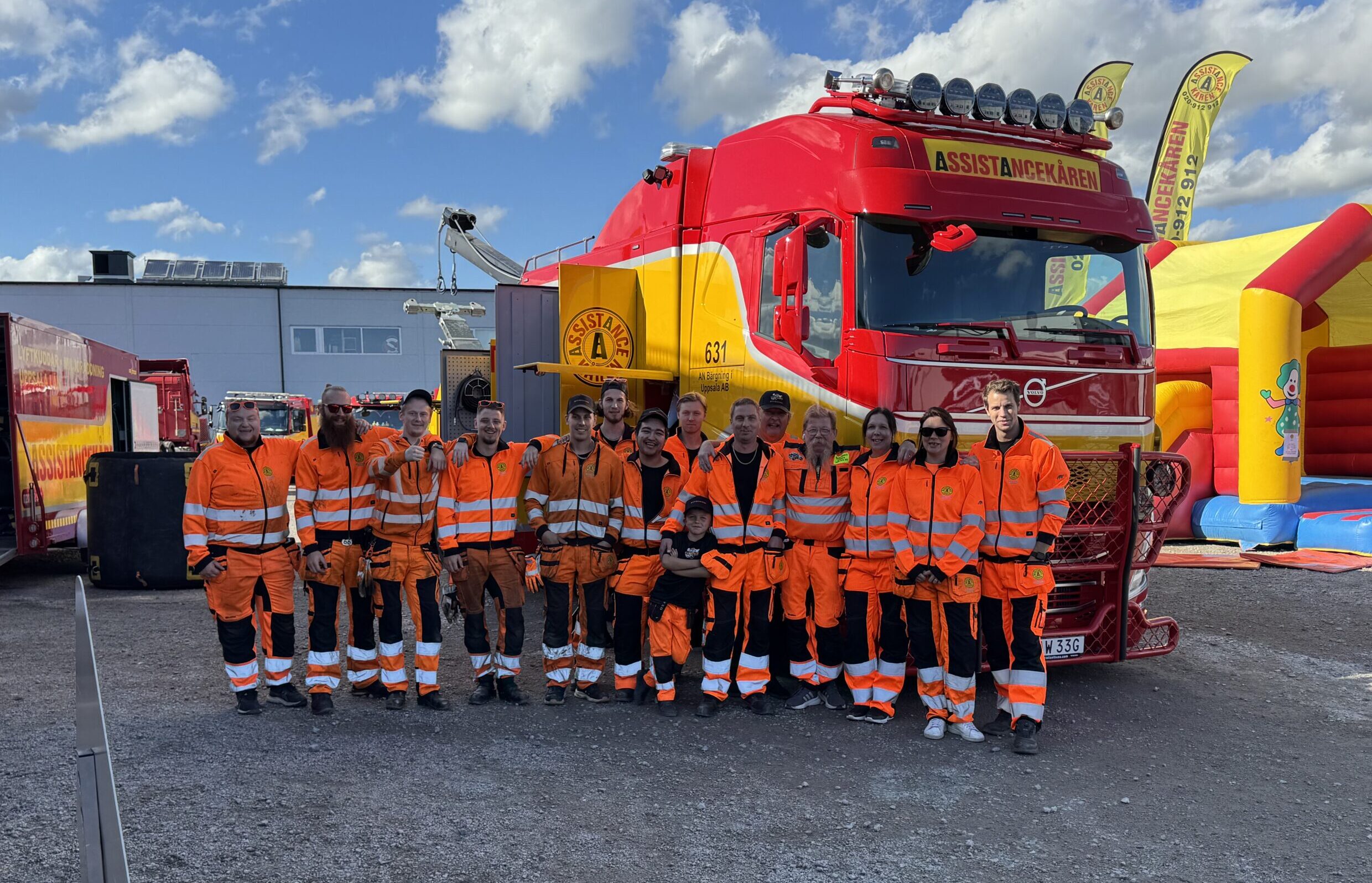 En grupp personer i orange arbetskläder från Assistanskåren står tillsammans framför en stor röd bärgningsbil. I bakgrunden syns en byggnad och en uppblåsbar lekattraktion under en blå himmel.