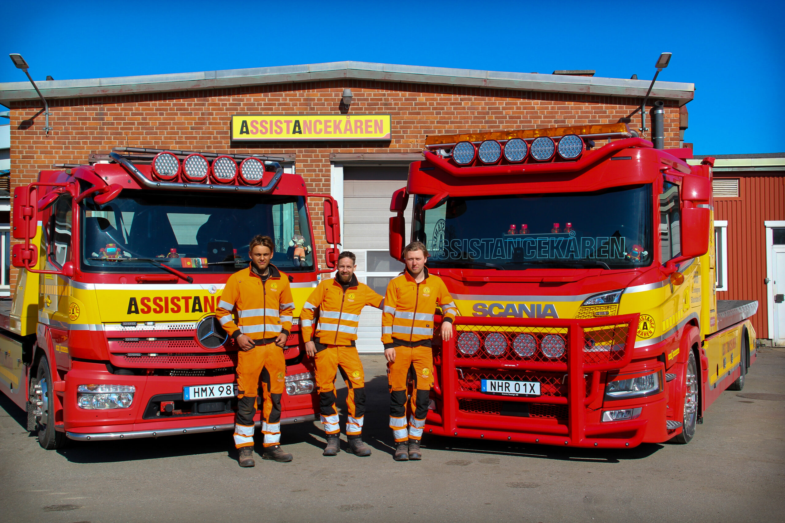 Tre medarbetare i orange arbetskläder står framför två röda bärgningsbilar från Assistancekåren Östhammar, parkerade utanför en garagebyggnad med skylten ”Assistancekåren”.