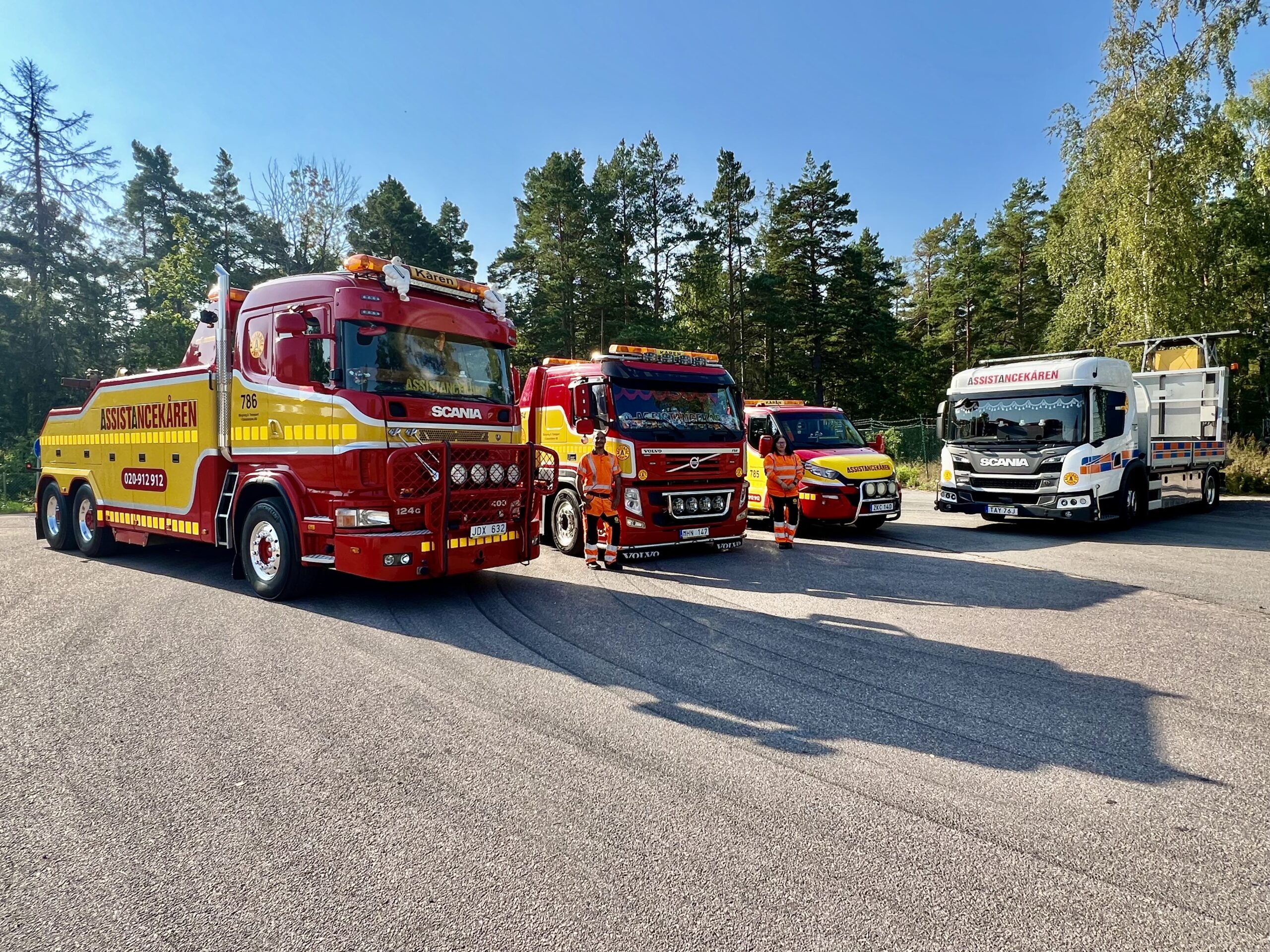 Fyra olika bärgningsfordon från Assistancekåren Oskarshamn står uppradade på en parkeringsplats, omgivna av skog. Fordonen inkluderar en tungbärgare, en medelstor bärgningsbil, en vägassistansbil och ett specialfordon för olyckshantering. Personal i uniform står stolt framför sina fordon, redo för alla typer av bärgningsuppdrag.