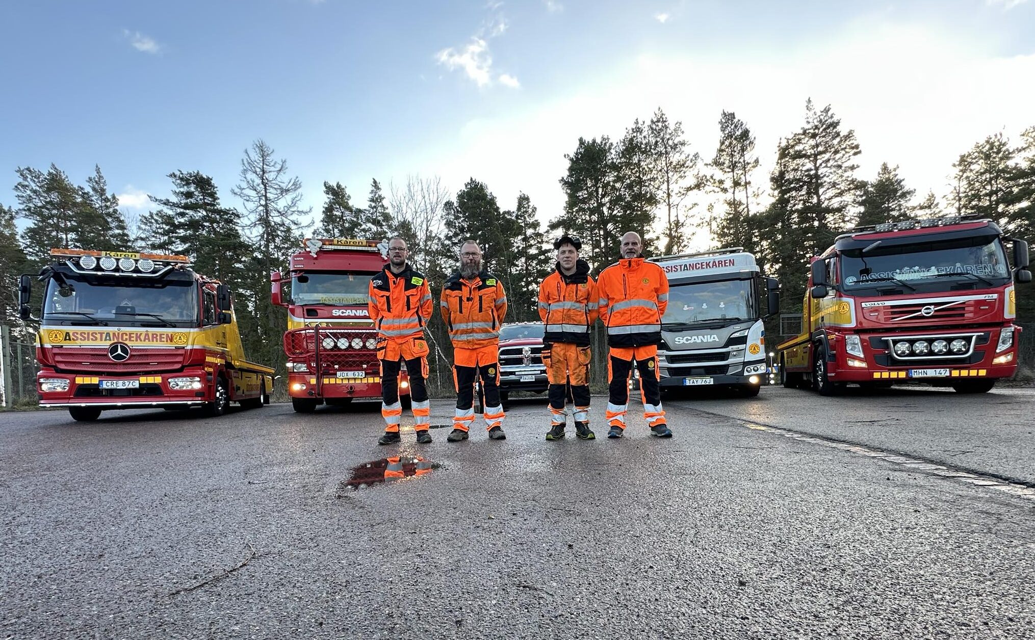 Fyra medarbetare från Assistancekåren Oskarshamn står framför flera röda och gula bärgningsbilar på en asfalterad yta med skog i bakgrunden.