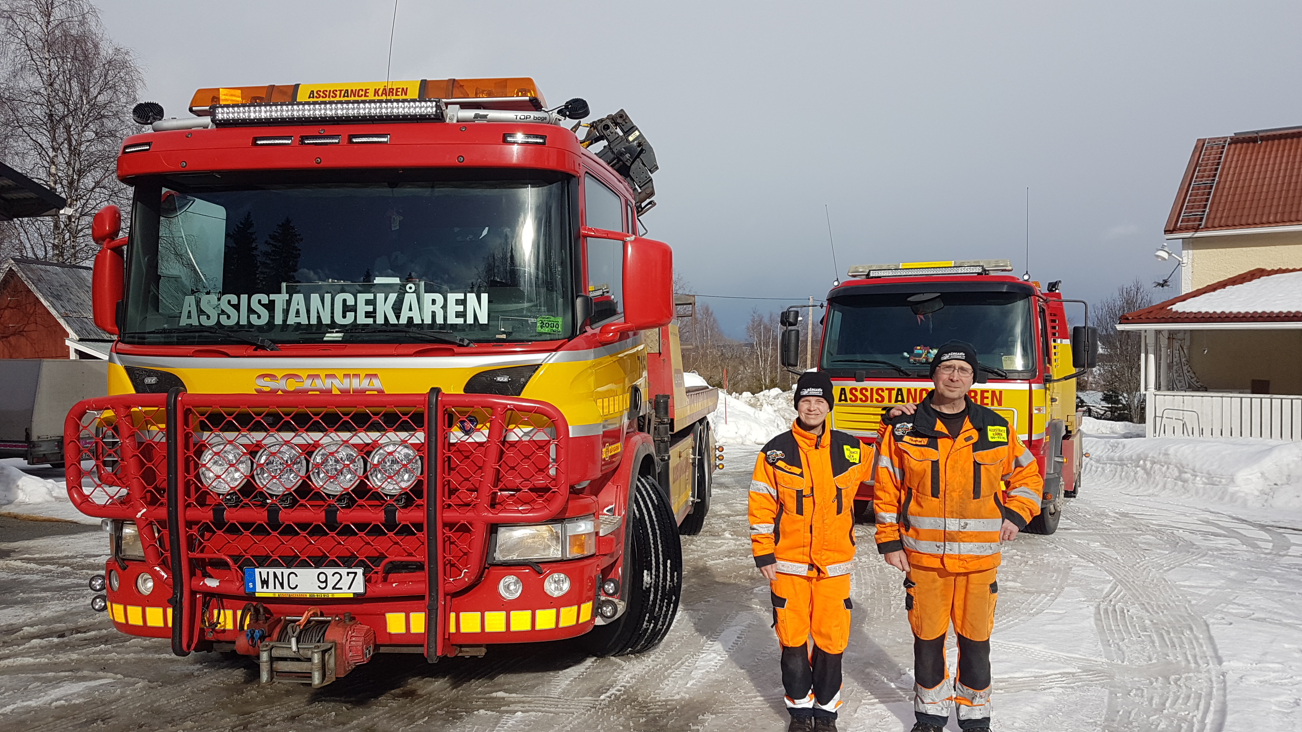 Två medarbetare från Assistancekåren Strömsund, iklädda orange arbetskläder och svarta mössor med Assistancekårens logotyp, står framför två röda och gula bärgningsbilar av märket Scania. Det är vinter, och marken är täckt av snö. Bilarna är utrustade med extraljus och varselutrustning, och "Assistancekåren" syns tydligt på fronten av fordonen.