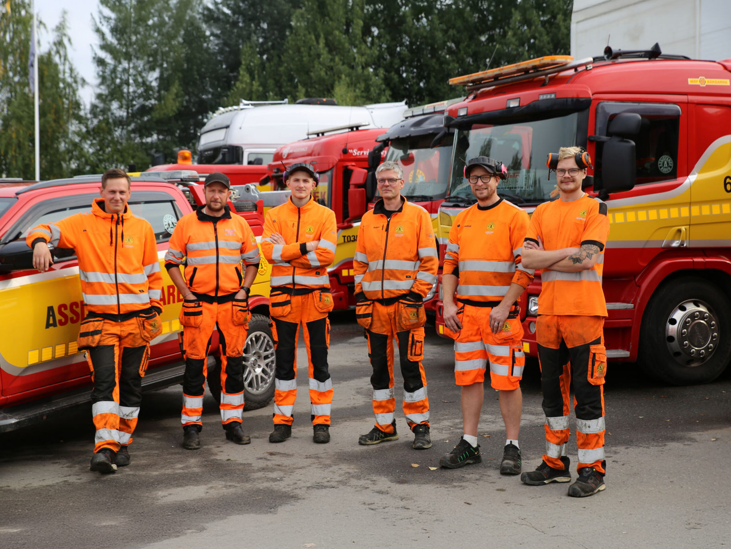 Sex medarbetare i orange arbetskläder står framför röda och gula bärgningsfordon från Assistancekåren på en uppställningsplats.