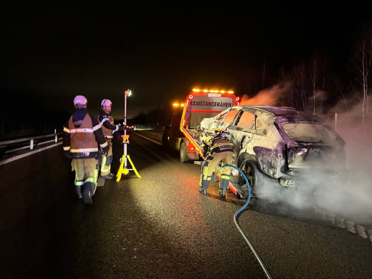 Räddningspersonal arbetar vid en hybridbil som brunnit under färd. Bilen, som är svårt brandskadad i bakdelen, står på vägbanan medan rök stiger upp från resterna. I bakgrunden syns Assistancekårens bärgningsfordon med varningsbelysning, och en upplyst arbetslampa lyser upp platsen. Situationen visar på de utmaningar och säkerhetsrisker som följer med moderna el- och hybridbilar vid bränder.