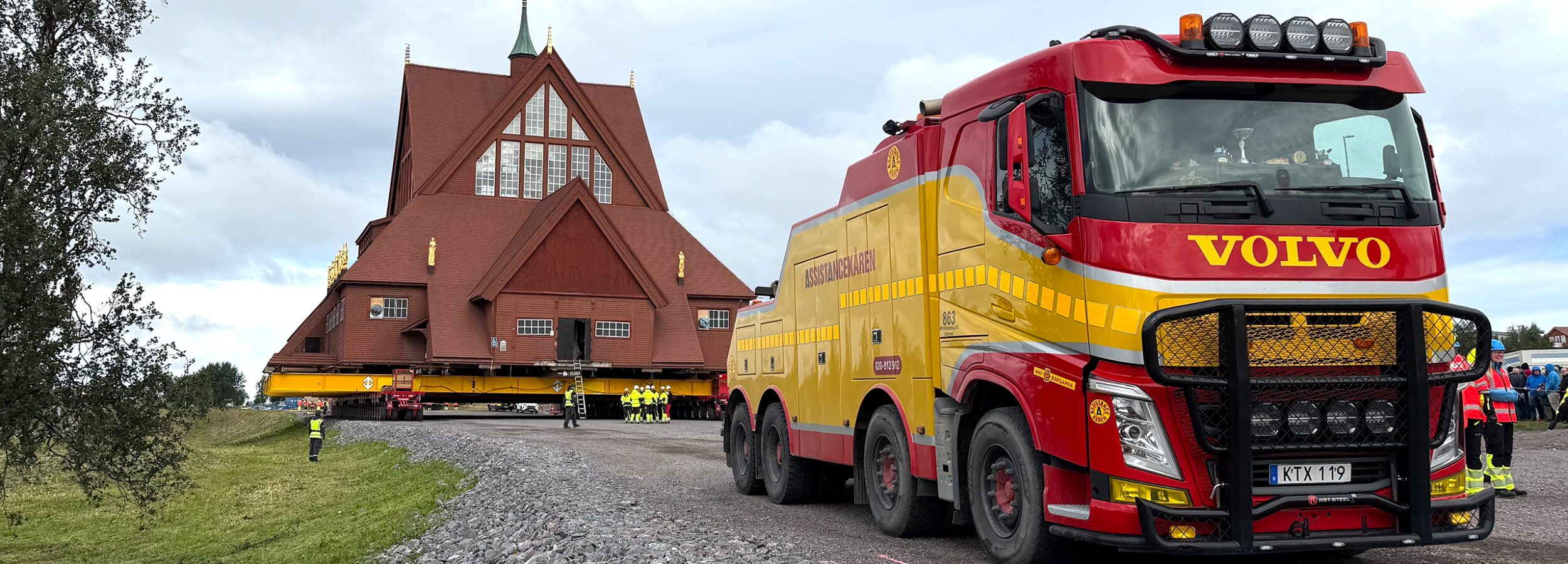 Kiruna kyrka på en stor trailer under transport, med Assistancekårens gula och röda lastbil i förgrunden. Flera personer i varselkläder står runt kyrkan och övervakar flytten på en grusväg.