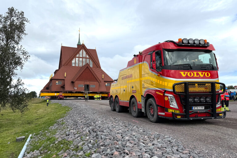 Kiruna kyrka på en stor trailer under transport, med Assistancekårens gula och röda lastbil i förgrunden. Flera personer i varselkläder står runt kyrkan och övervakar flytten på en grusväg.