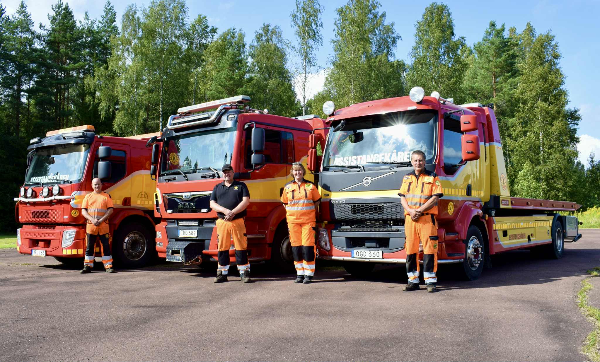 Fyra bärgare i arbetskläder står framför tre bärgningsfordon från Assistancekåren i Deje och Hagfors. De står på asfalterad yta, med skog i bakgrunden.