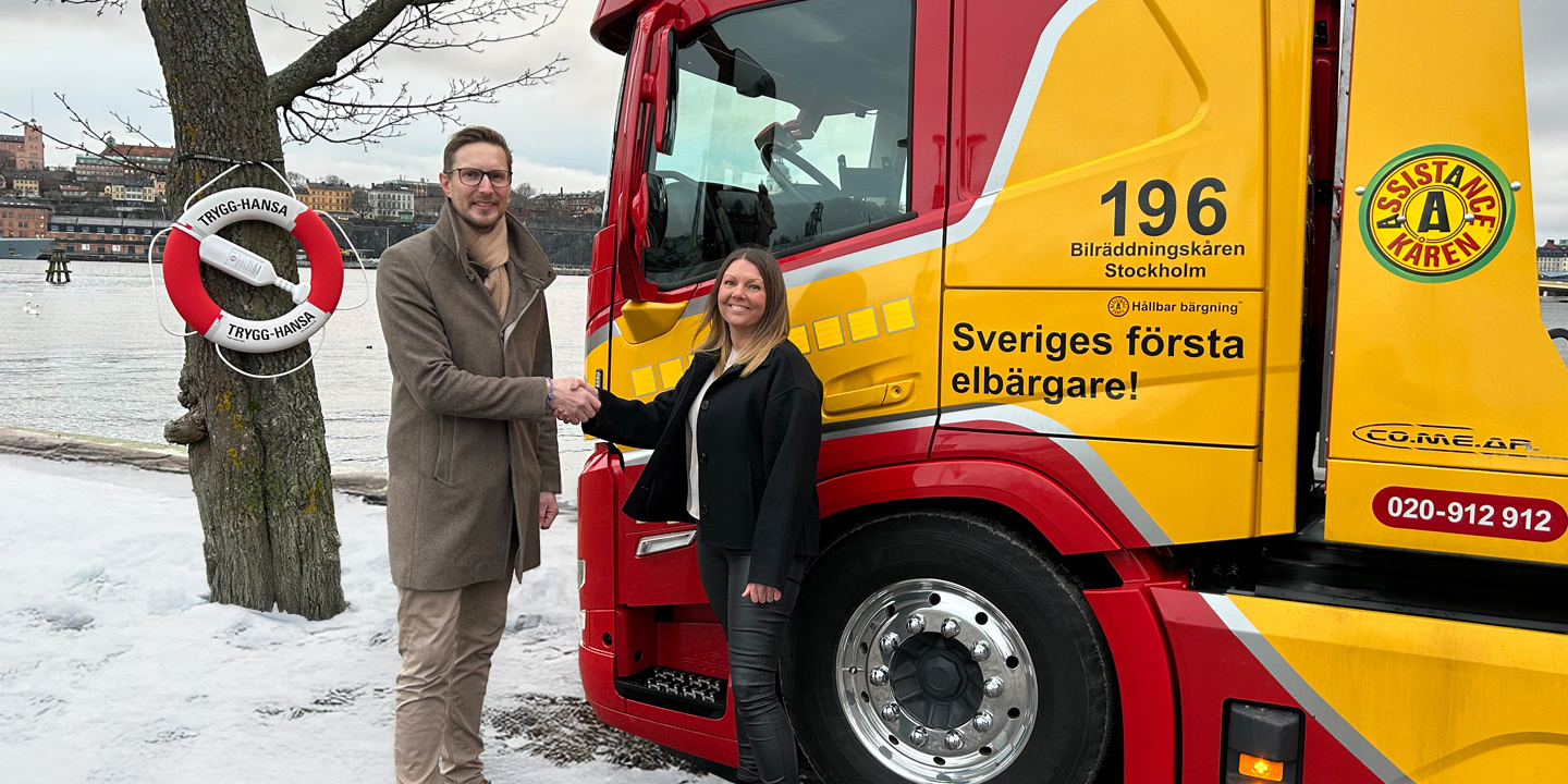 Svante Hedbring från Trygg-Hansa och Linda Gyllensvärd, vd för Assistancekåren, skakar hand framför en Assistancekåren-bärgningsbil i samband med det nya avtalet för bärgning och vägassistans, med en Trygg-Hansa-boj i bakgrunden.