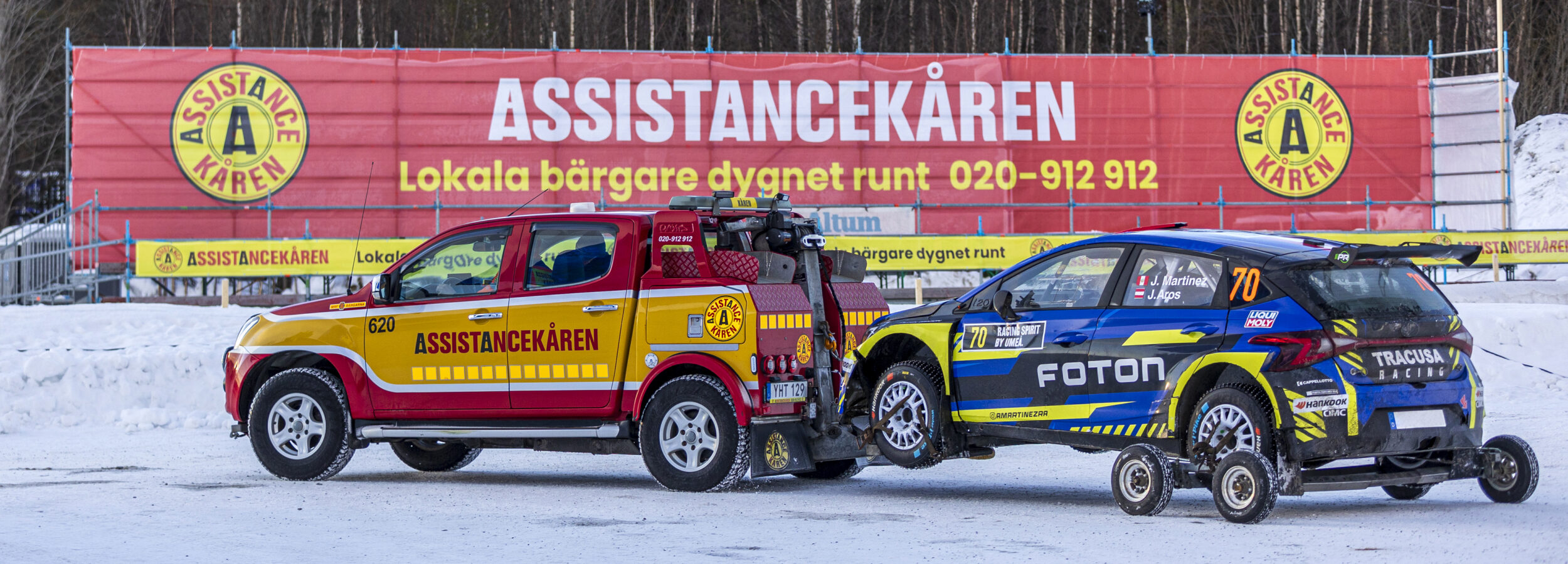 En bärgningsbil från Assistancekåren bogserar en rallybil på snöunderlag vid Rally Sweden. I bakgrunden syns Assistancekårens tillgänglighetsanpassade läktare på Red Barn Arena med tydlig Assistancekåren-skyltning.