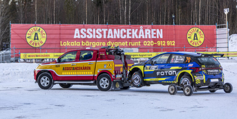 En bärgningsbil från Assistancekåren bogserar en rallybil på snöunderlag vid Rally Sweden. I bakgrunden syns Assistancekårens tillgänglighetsanpassade läktare på Red Barn Arena med tydlig Assistancekåren-skyltning.