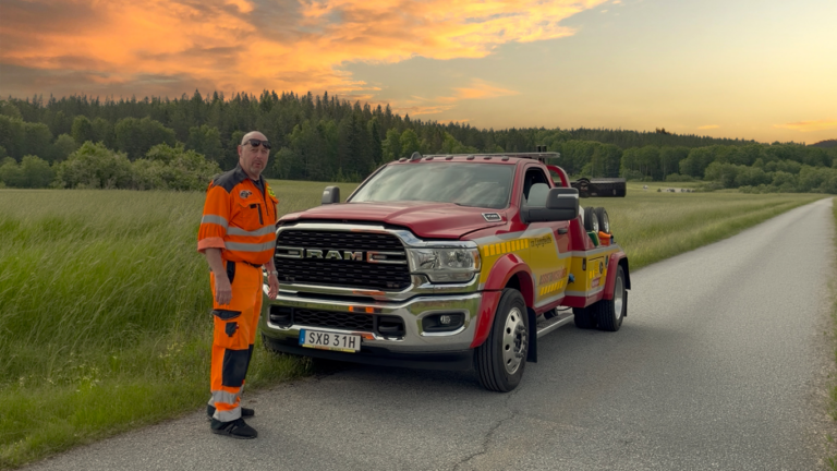 Bärgningsbil från Assistancekåren står parkerad på en smal landsväg vid en äng i solnedgång, med en bärgare i orange varselkläder bredvid fordonet.