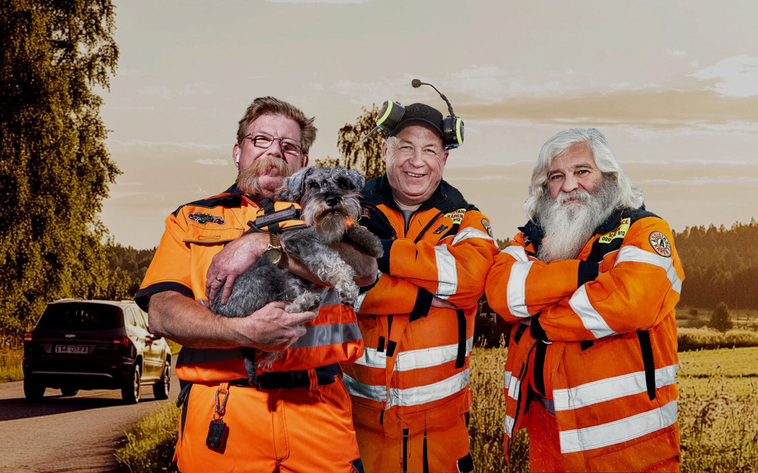 Bärgar-Berra, Stefan och Kent från Assistancekåren står tillsammans i sina orange arbetskläder vid en landsväg. Bärgar-Berra håller en liten grå hund i famnen. En bil syns i bakgrunden och bilden illustrerar Assistancekårens bärgning och vägassistans i en trygg och välkomnande miljö.