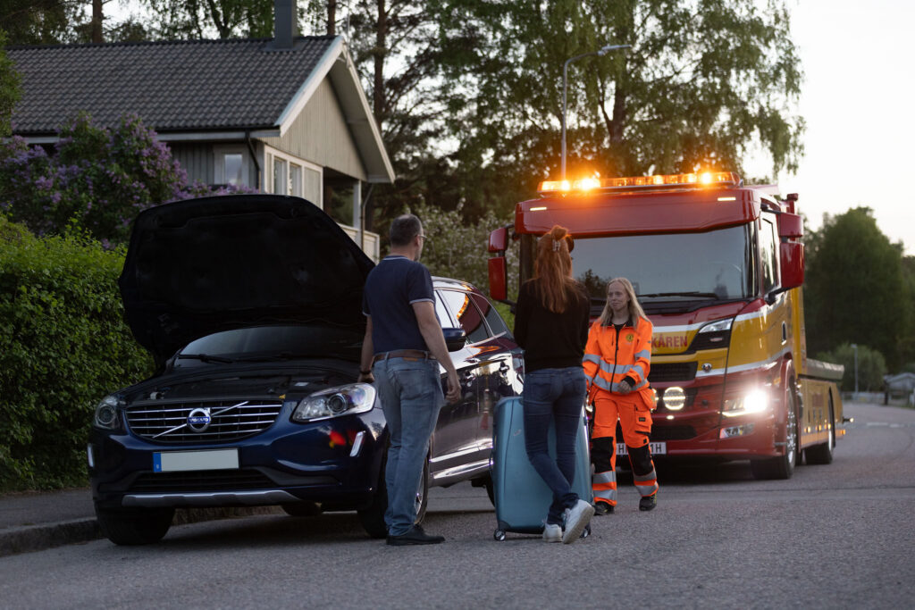 En medarbetare från Assistancekåren hjälper två personer vid en bil med öppen motorhuv på en villagata. Bärgningsbilen står bakom med varningsljus påslagna medan resenärerna väntar bredvid bilen med en resväska.