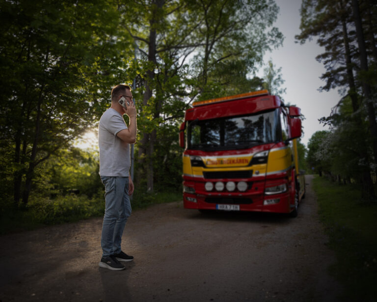 Person står på en grusväg i skogsmiljö och pratar i mobiltelefon. En röd och gul bärgningsbil från Assistancekåren står parkerad framför personen. Solen lyser genom träden i bakgrunden och vägen ser fuktig ut efter regn.