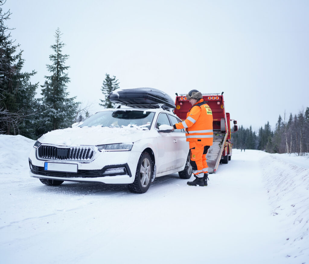 En bärgare från Assistancekåren hjälper en bilist med en snötäckt vit personbil på en vinterväg. Bakom bilen står en röd bärgningsbil redo för transport.